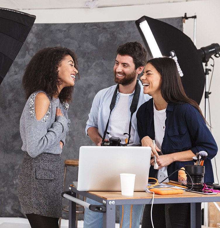 Three colleagues laughing together, with one man holding a camera and two women smiling in the background near a desk with a laptop and a large light source behind them.