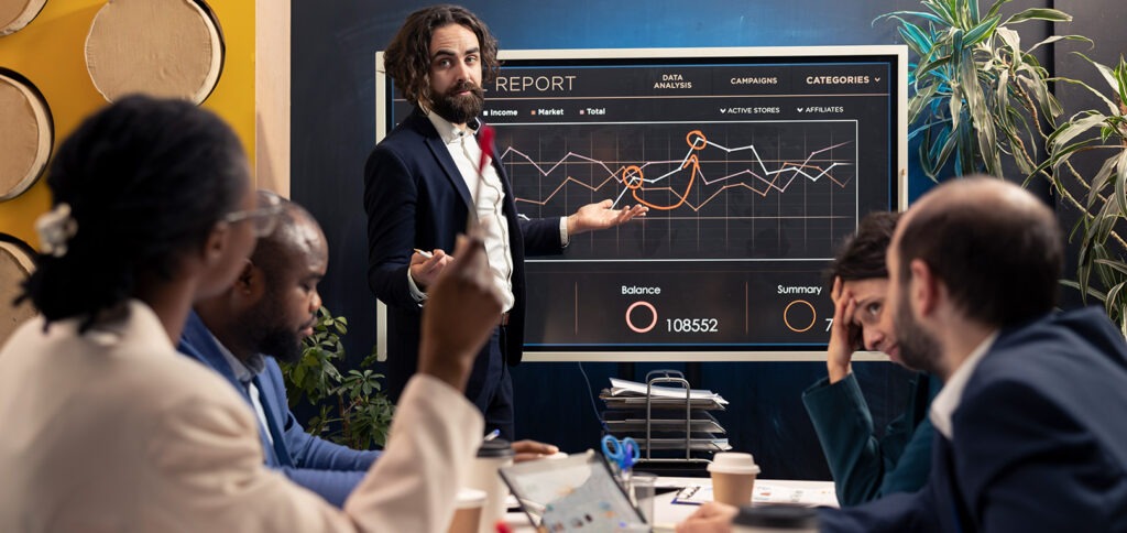 A group of people in a meeting with one person presenting a graph showing ups and downs, while another employee looks slightly disappointed.