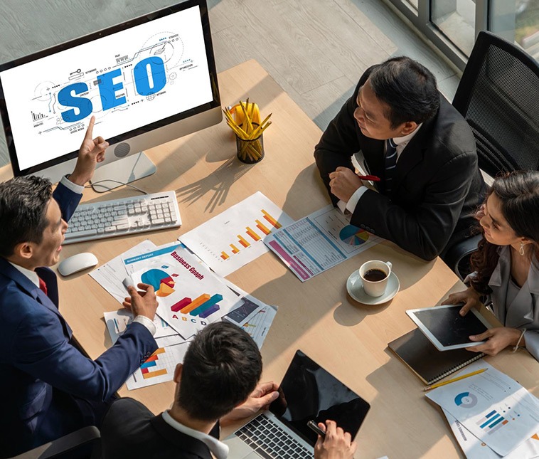 Four employees sitting around a table during a meeting, with one pointing at a laptop displaying an SEO screen."