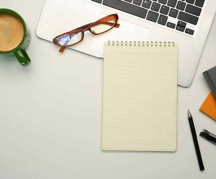 A work desk with a cup of coffee, a laptop, a notepad, a pen, and a pair of glasses.