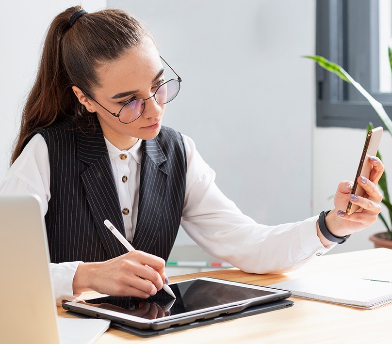 A woman holding her phone while writing on her iPad in a work environment.