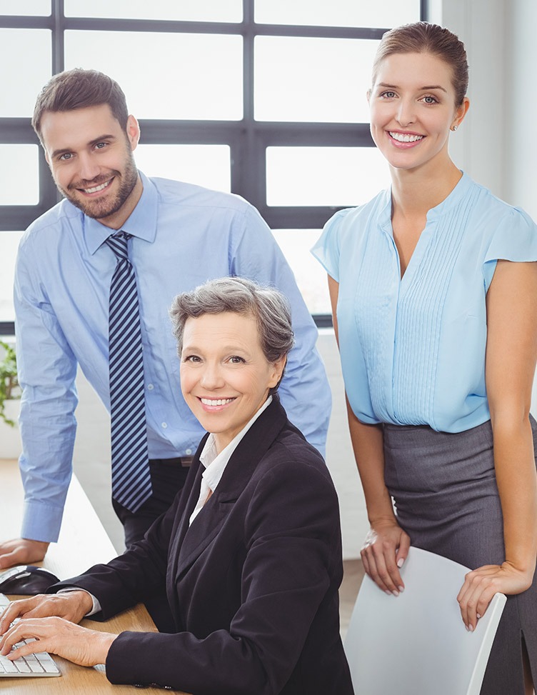 Three professionals in an office setting, with one sitting, one standing, and one leaning on the table, all smiling.